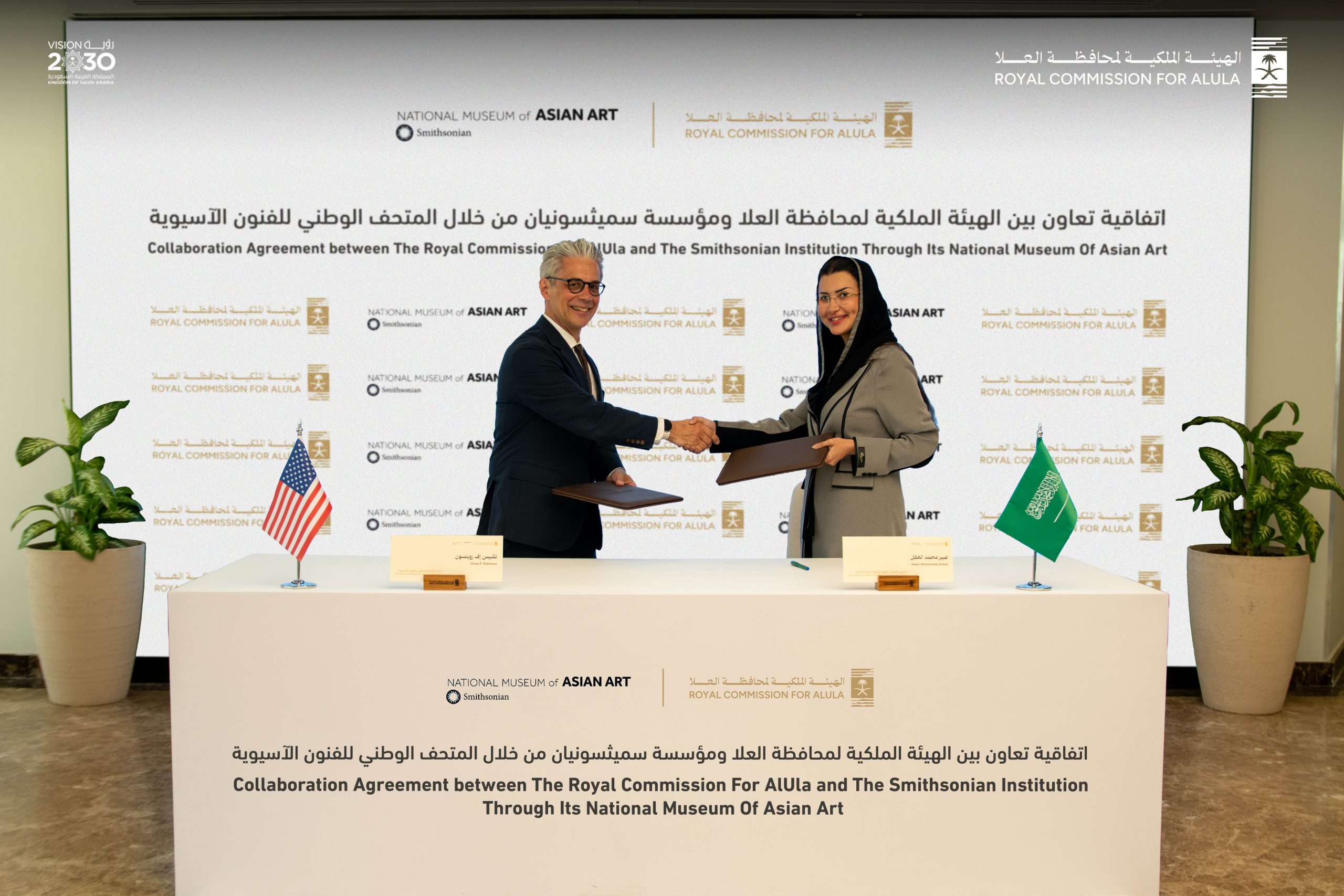 A man and a woman standing behind a white table shake hands and smile at the camera at a ceremonial signing
