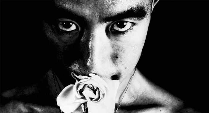 Black and white film still of a face with intense eyes staring at the camera, while the subject holdsa a white flower in their mouth.