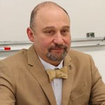 Pavel Lurje in a light brown suit and bowtie, in front of a whiteboard.