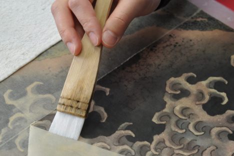 a hand holding a brush against a piece of paper laid over a paint showing stylized waves